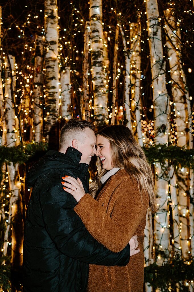 A couple embrace and smile at one another while standing in front of a structure of birch trees and lights inside of the Jack Frost Market