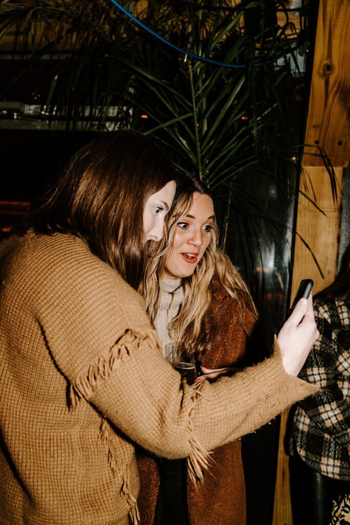 A woman and her friend facetime another friend on a phone while inside of a bar