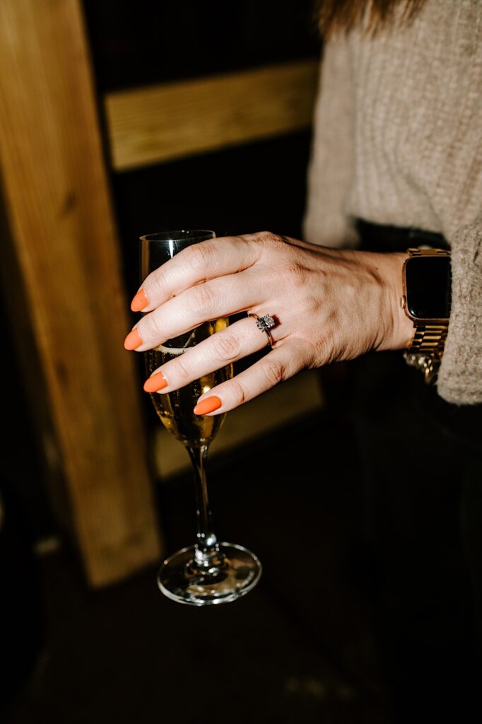 Close up photo of a woman's hand that shows off her engagement ring as she holds a glass of champagne