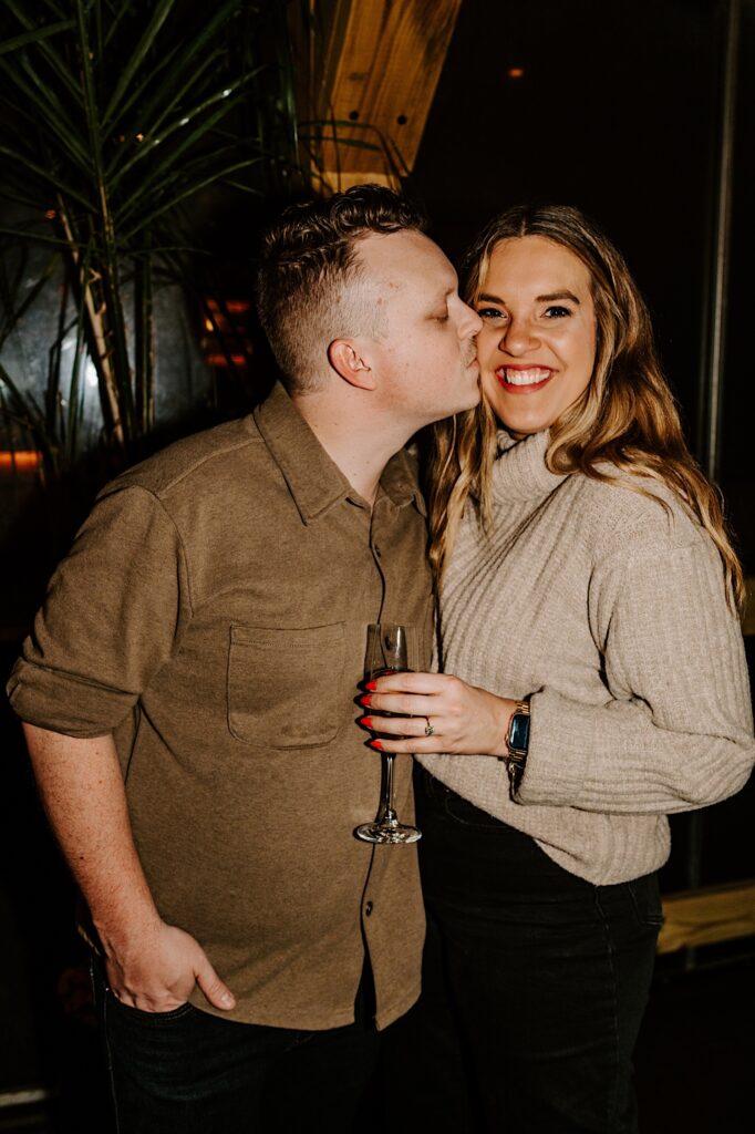 A woman smiles at the camera while being kissed by a man on the cheek inside of a bar in Chicago as the woman shows off her engagement ring to the camera