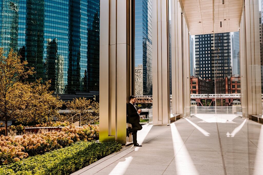 A groom stands outside of his wedding venue, the Voco Hotel in Chicago, and leans against a pillar on a sunny day