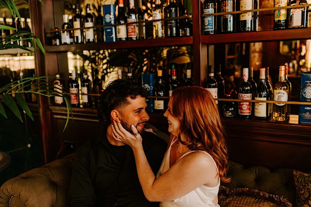 A couple smile at one another while sitting on a couch in a Chicago bar as they have their engagement photos taken