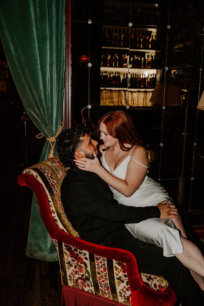 A woman sits on a man's lap while he sits in a red floral chair in a bar of Chicago