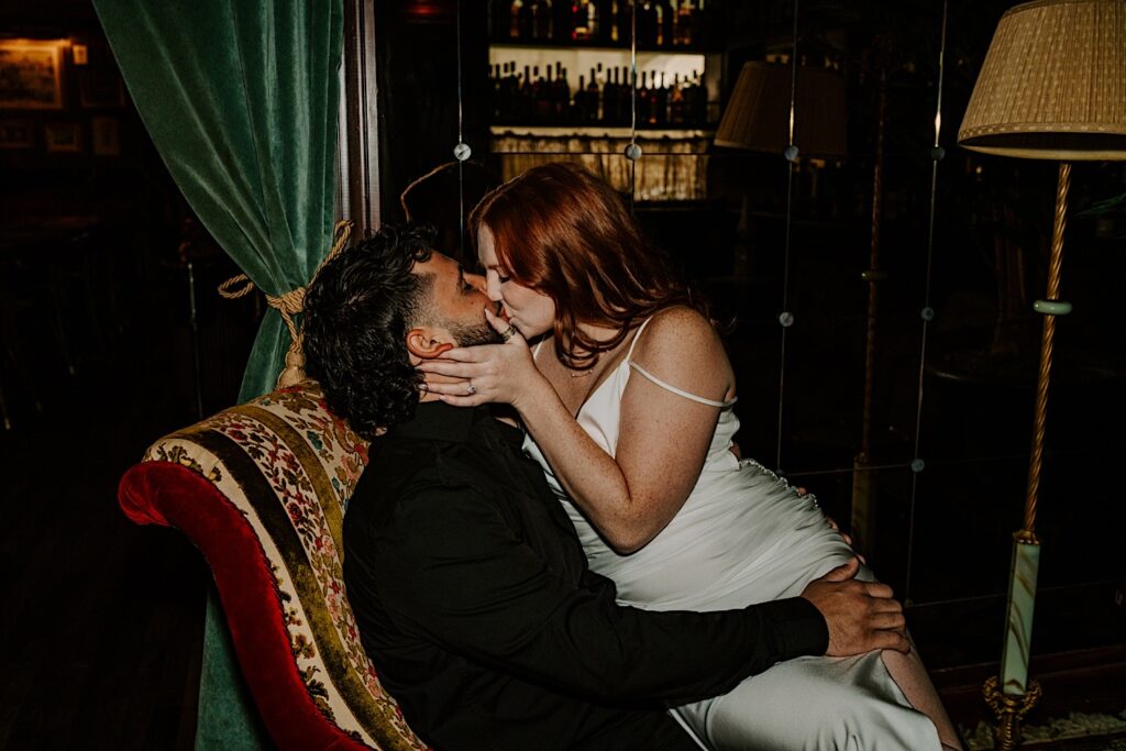 A woman kisses a man as she sits on his lap while he sits in a red floral chair in a Chicago bar