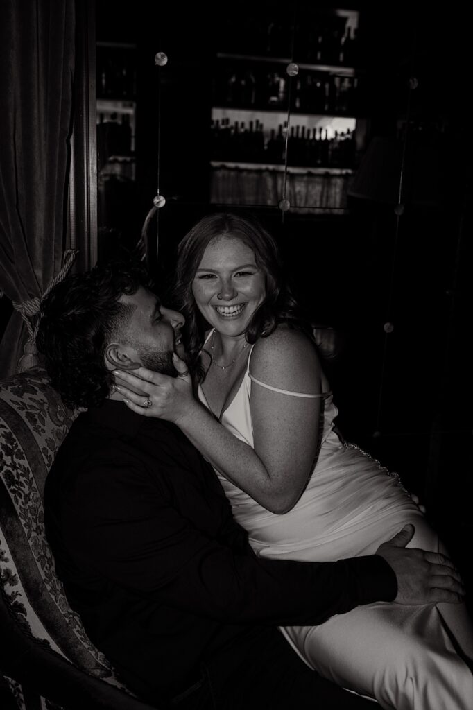 Black and white photo of a woman smiling at the camera while sitting in a man's lap as he smiles up at her