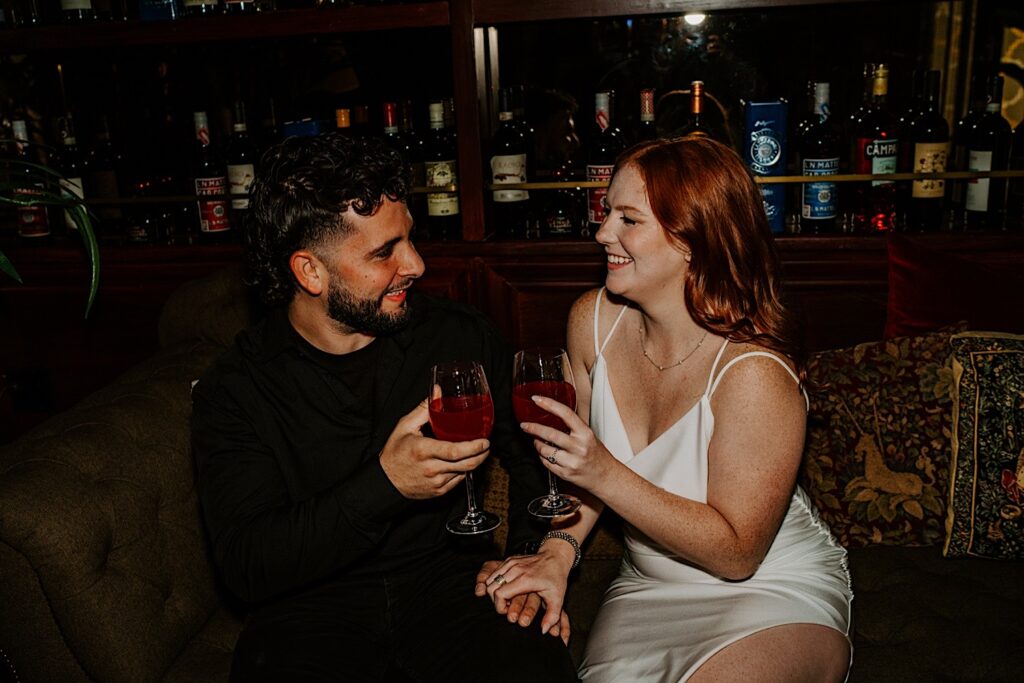 A couple sit together on a couch in a Chicago bar and smile at each other as they hold glasses of red wine