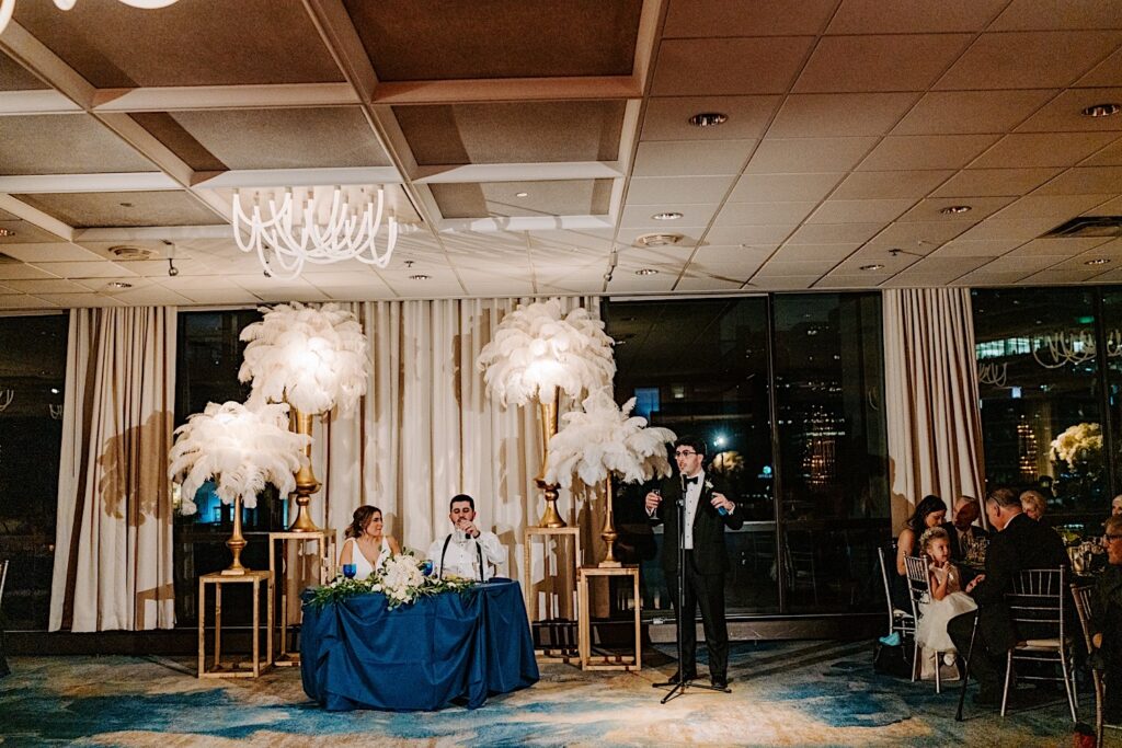A bride and groom sit at their sweetheart table as a groomsman gives a speech next to them while in the indoor reception space of their wedding venue, the Voco Hotel in Chicago