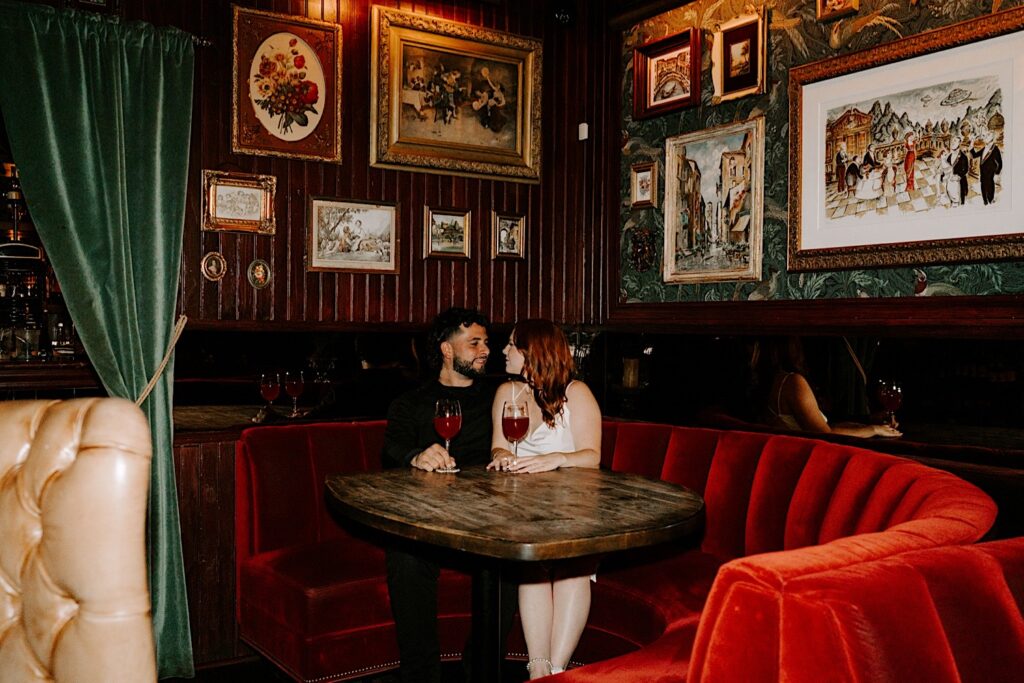 A couple sit in a red velvet booth of a Chicago bar and smile at one another while having their engagement photos taken