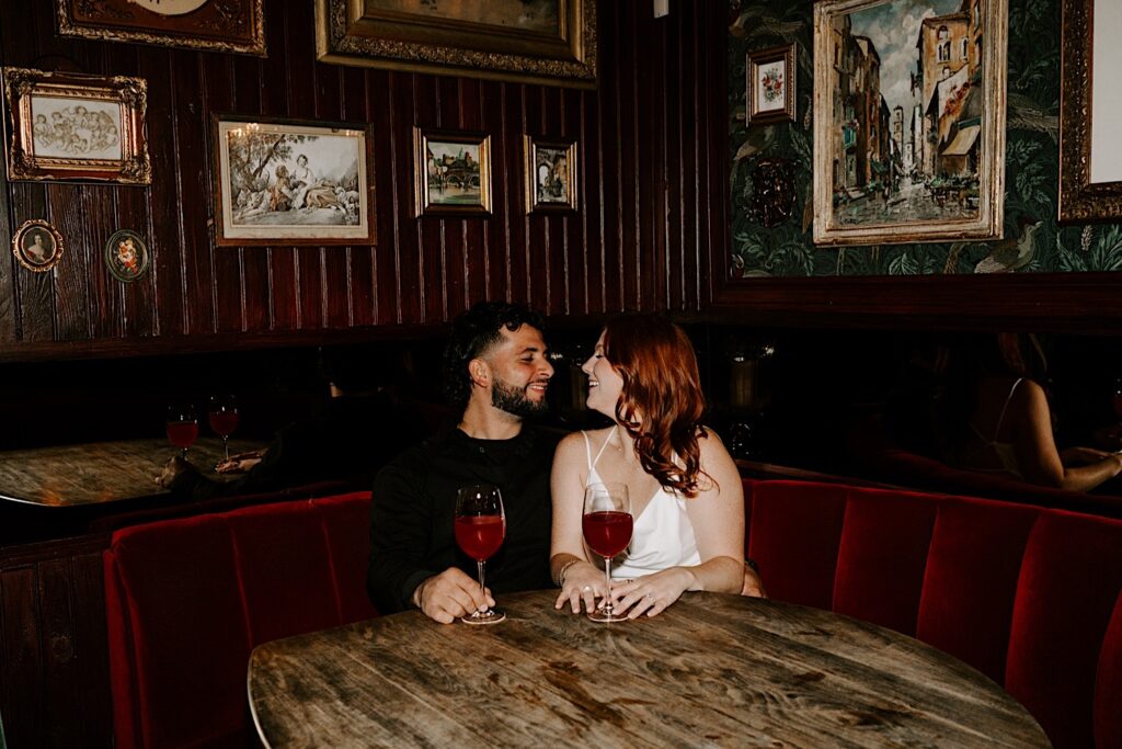 A couple smile at one another while drinking at a bar in Chicago together as they sit in a red velvet booth
