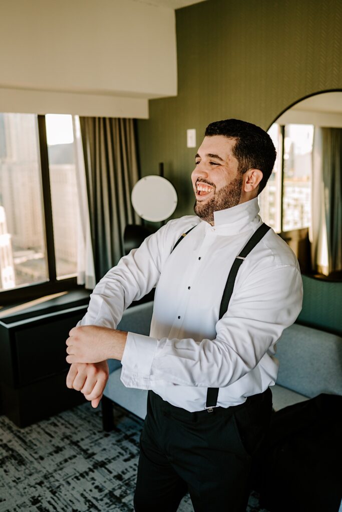 A groom smiles while getting dressed for his wedding day while inside his hotel room