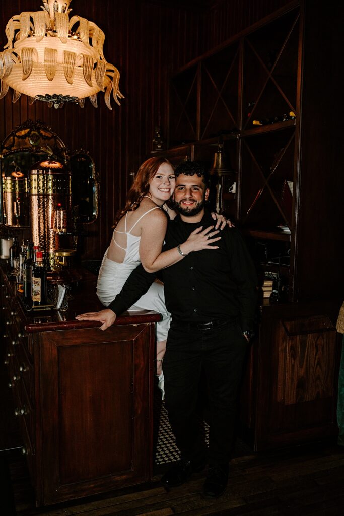 A couple smile at the camera, the man standing while the woman sits on a bar behind him and hugs him