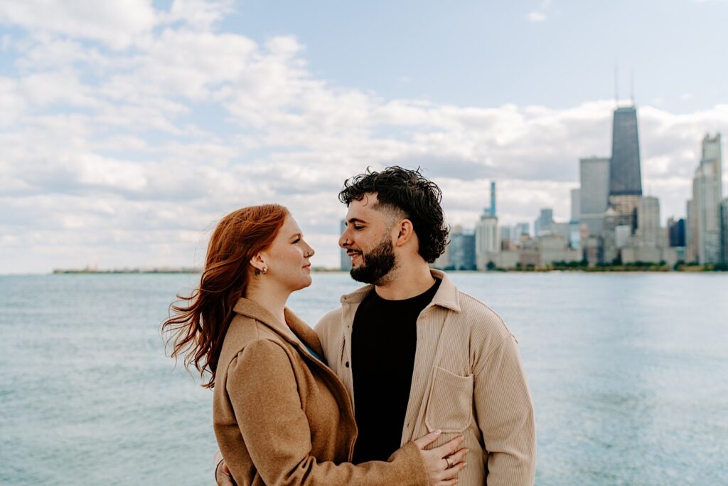 A couple smile at one another while having their engagement photos taken at Chicago's North Avenue Beach. Behind them is Lake Michigan and the city's skyline