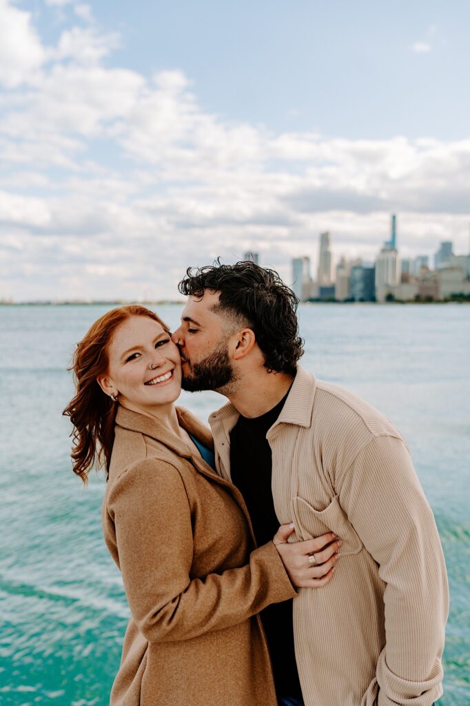 A woman smiles at the camera while a man kisses her cheek as they stand in front of Lake Michigan and the Chicago skyline