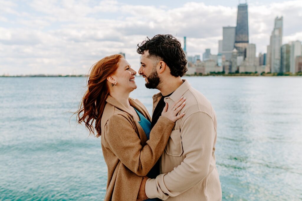 A couple embrace and smile at each other while having their engagement photos taken at North Avenue Beach in Chicago, behind them is Lake Michigan and the city skyline