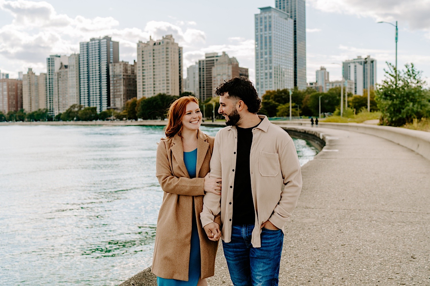 A couple smile at one another while holding hands and walking along North Avenue Beach in Chicago as they have their engagement photos taken