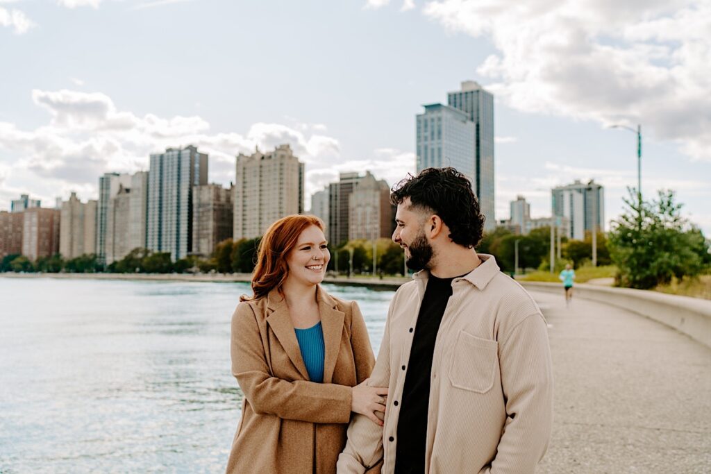 A couple smile at one another while the woman holds the man's arm as they have their engagement photos taken at North Avenue Beach in Chicago. Behind them is Lake Michigan and the city's skyline