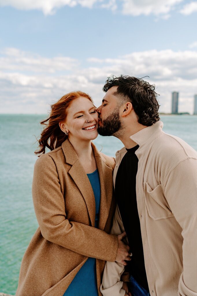 A woman smiles as a man kisses her on the cheek, behind them is Lake Michigan and a few skyscrapers of Chicago