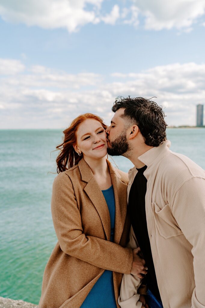 A woman smiles as a man kisses her on the cheek while they stand in front of Lake Michigan