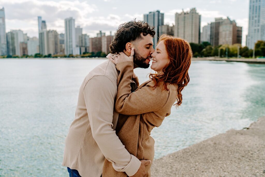 A woman smiles as a man leans in to kiss her while they have their engagement photos taken at North Avenue Beach in Chicago with Lake Michigan and the city's skyline behind them