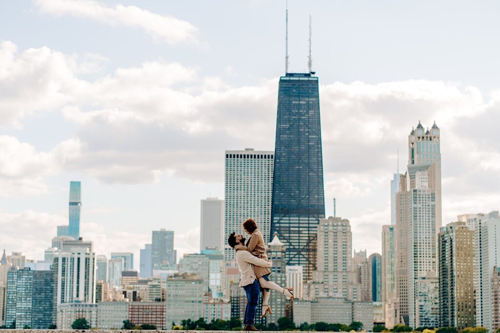 A man lifts a woman in the air in front of the buildings of Chicago while they have their engagement photos taken at North Avenue Beach