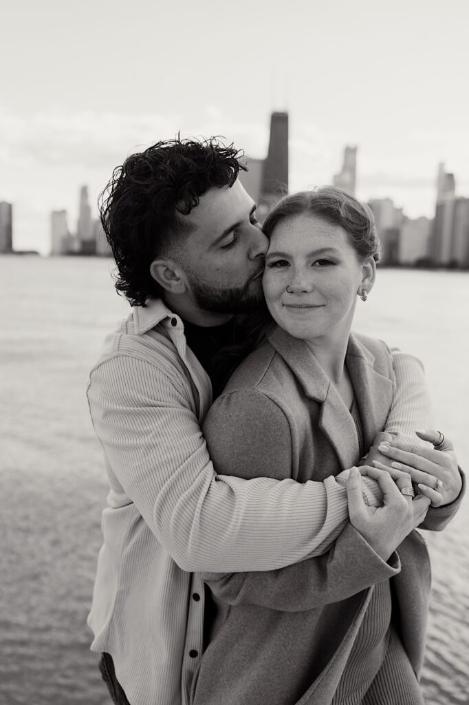 Black and white photo of a woman smiling at the camera as a man hugs her from behind and kisses her cheek, behind them is Lake Michigan and the Chicago skyline