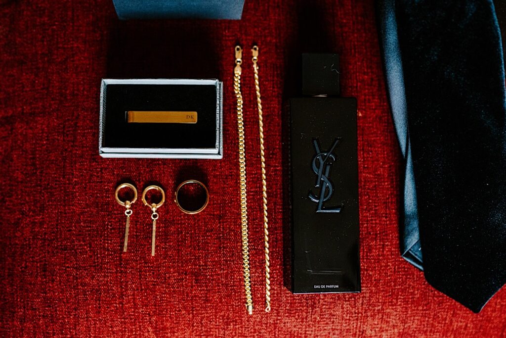 A wedding day flat lay consisting of cologne, rings, a tie, and more jewelry