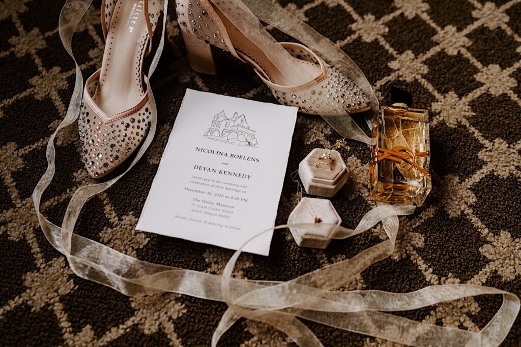 Wedding day flat lay consisting of rings, perfume, an invitation, and shoes all on a carpet