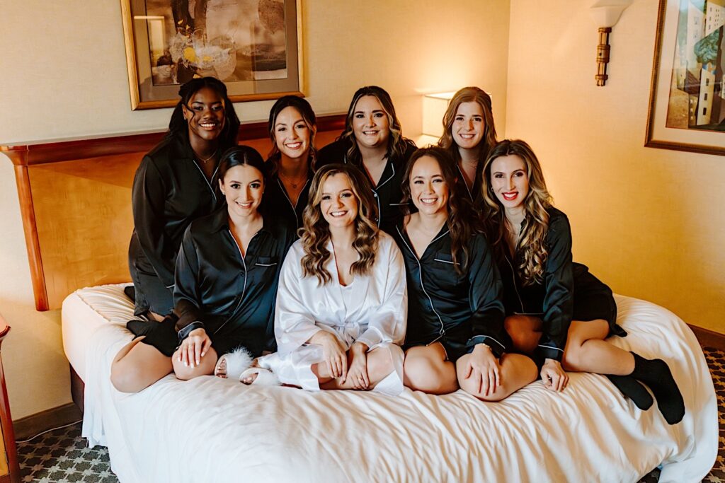 A bride sits on a hotel bed with her 7 bridesmaids as they all smile at the camera before getting ready for the wedding