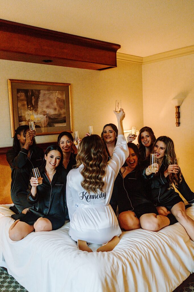 A bride sits on a bed with her 7 bridesmaids as they all raise glasses of champagne in the air, the bride faces away from the camera while the bridesmaids face towards her
