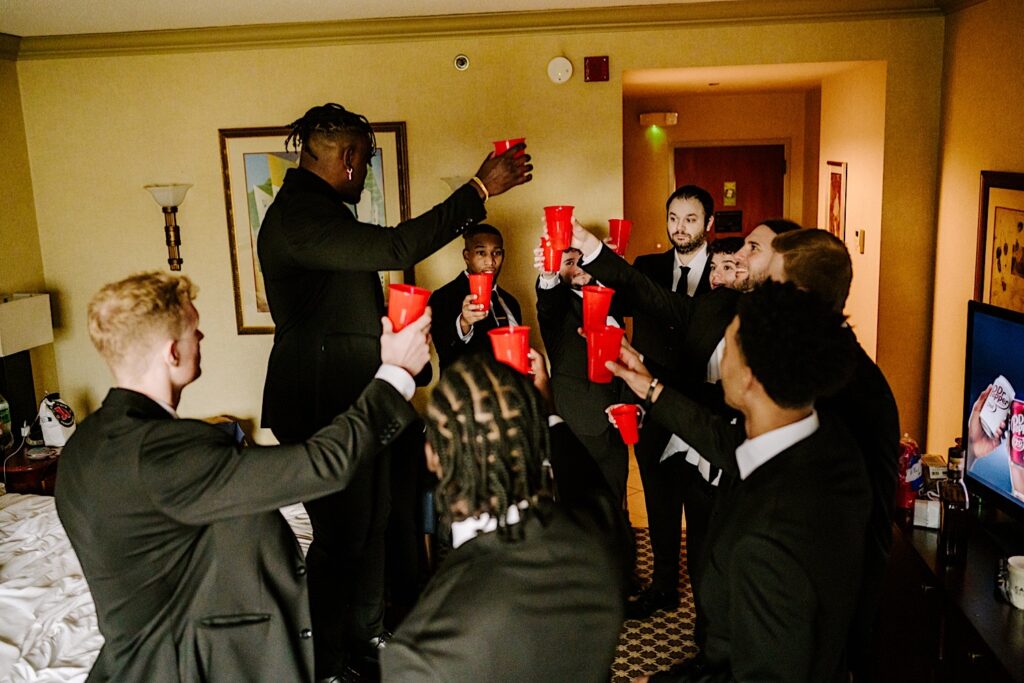 A groom stands in a hotel room with his groomsmen as they all raise plastic red cups in the air for a toast
