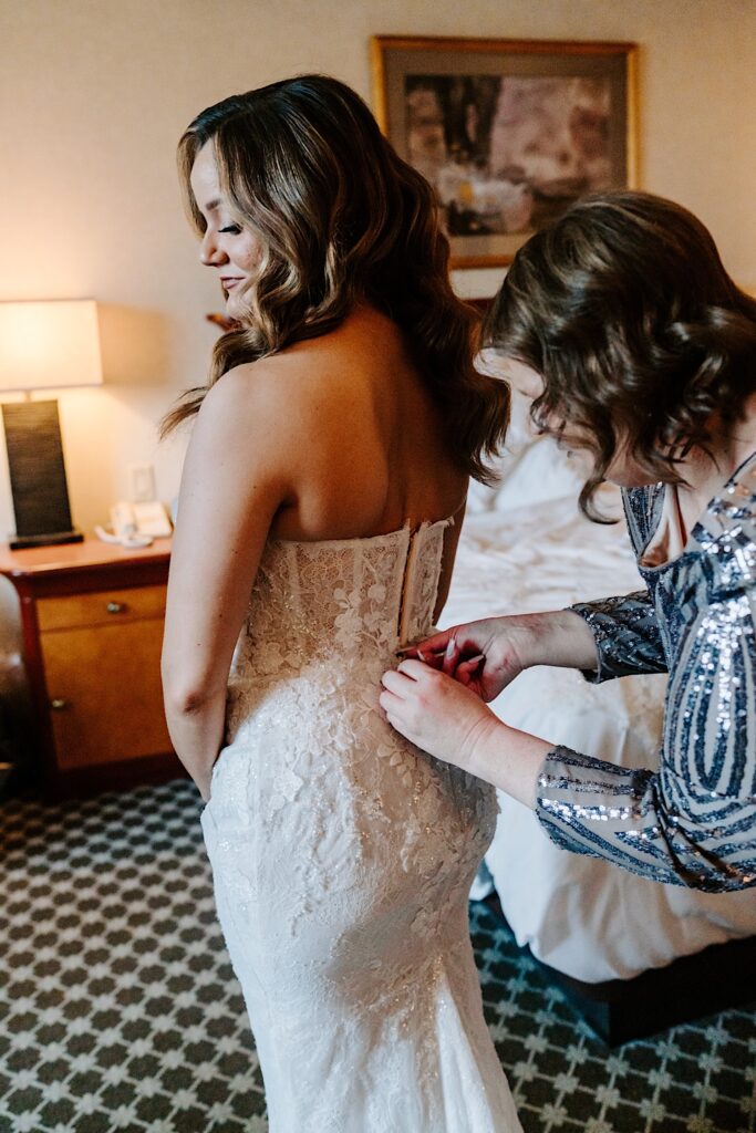A bride smiles as her mother buttons up the back of her wedding dress while getting ready in a hotel room