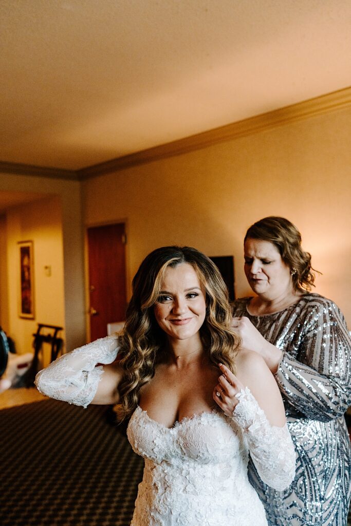 A bride smiles at the camera while her mother stands behind her and adjusts the back of her wedding dress