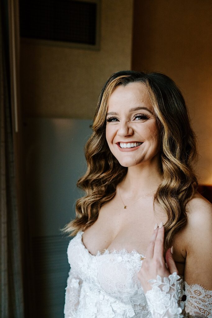 A bride in her wedding dress smiles at the camera while standing in front of a window of her hotel room
