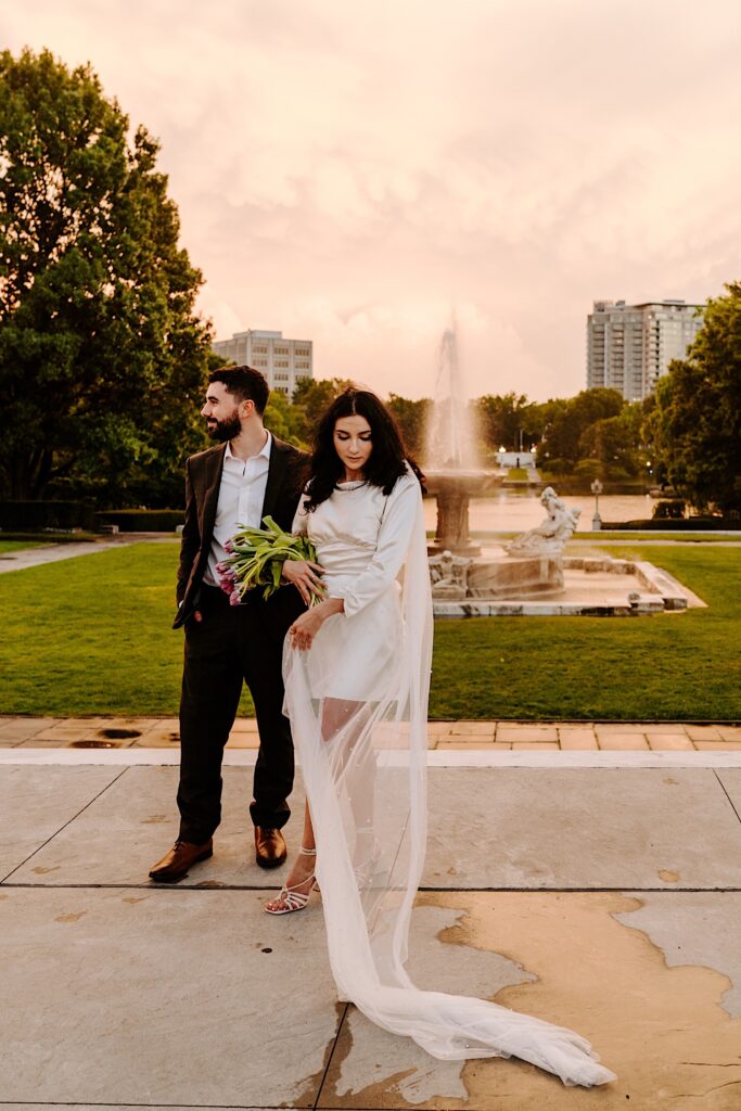 A bride and groom stand for a portrait photo in front of a fountain at sunset