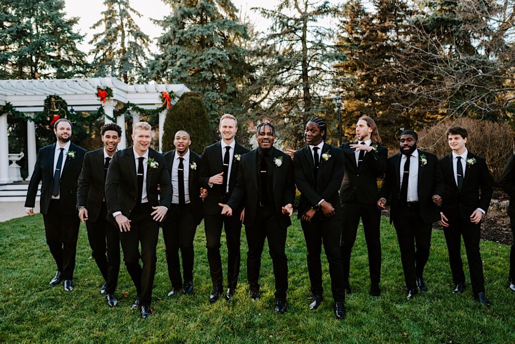 A groom walks with his 9 groomsmen towards the camera while taking wedding party photos outside of The Haley Mansion