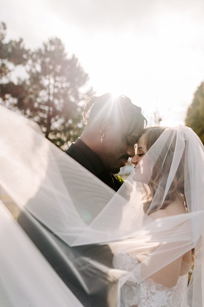 A bride and groom close their eyes and touch their heads together while the sun shines behind them