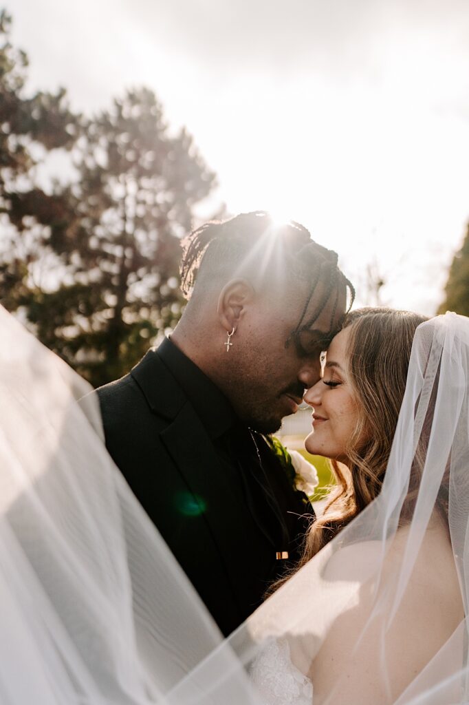 A bride and groom close their eyes and touch their heads together while the sun shines behind them