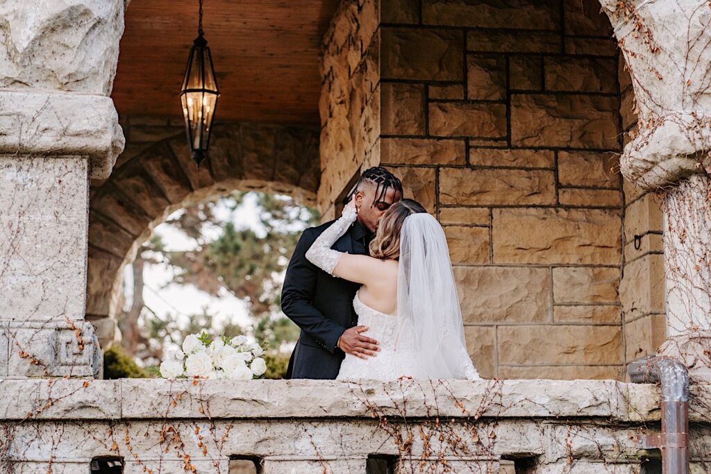 A bride and groom kiss one another while on a stone balcony outside of The Haley Mansion before their wedding ceremony there