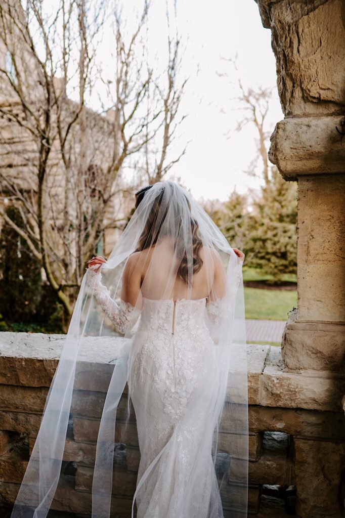 A bride plays with her veil while facing away from the camera and looking out over a stone balcony at The Haley Mansion