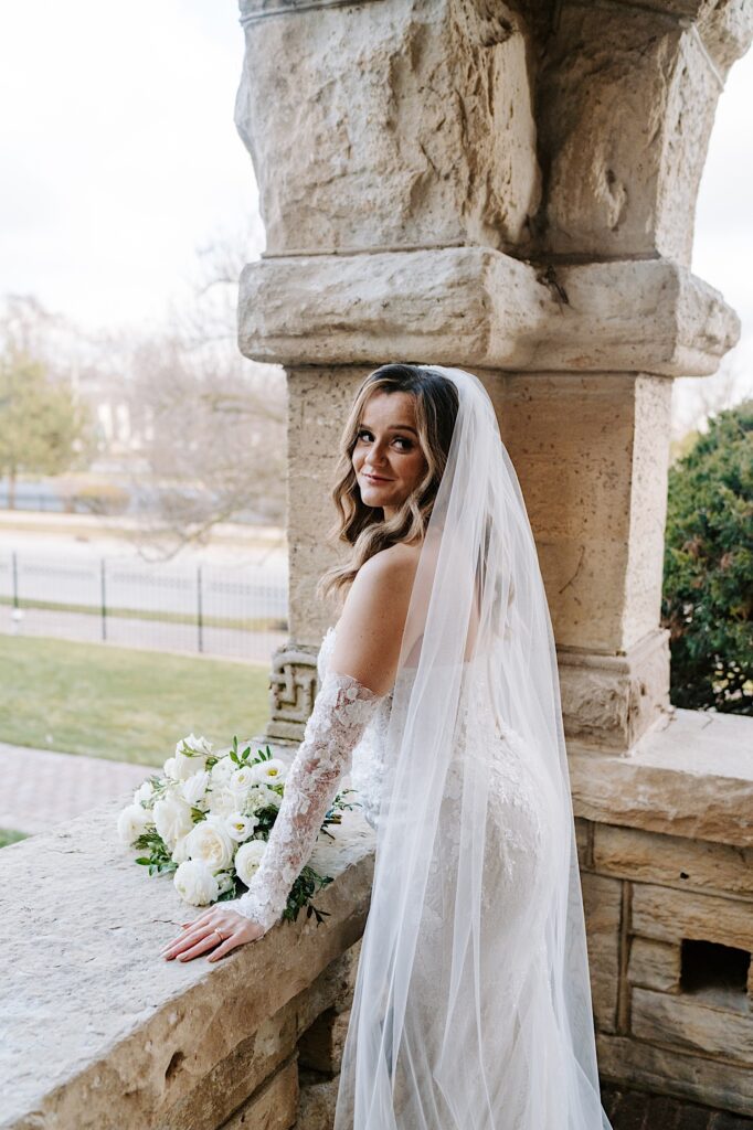 A bride leans against a stone balcony at The Haley Mansion and smiles at the camera over her shoulder
