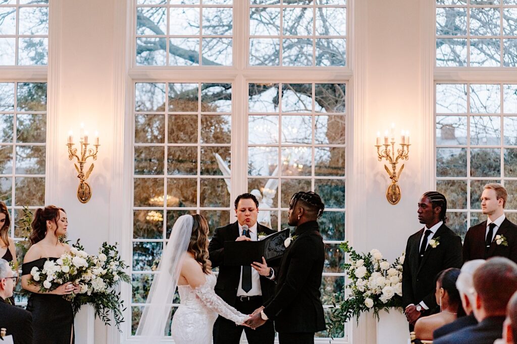 A bride and groom hold hands while their officiant speaks during their indoor wedding ceremony at The Haley Mansion