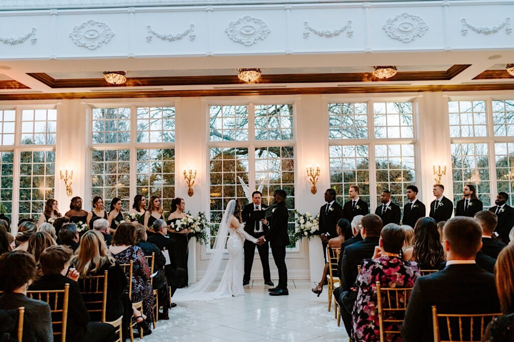 A bride and groom hold hands as their officiant speaks and guests watch during their wedding ceremony at The Haley Mansion