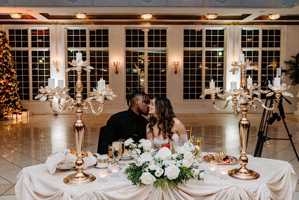 A bride and groom kiss one another while sitting at their sweetheart table during their wedding reception at The Haley Mansion