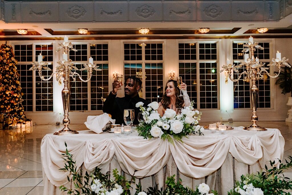 A bride and groom sit at their sweetheart table and raise their glasses during a speech at their indoor wedding reception in The Haley Mansion