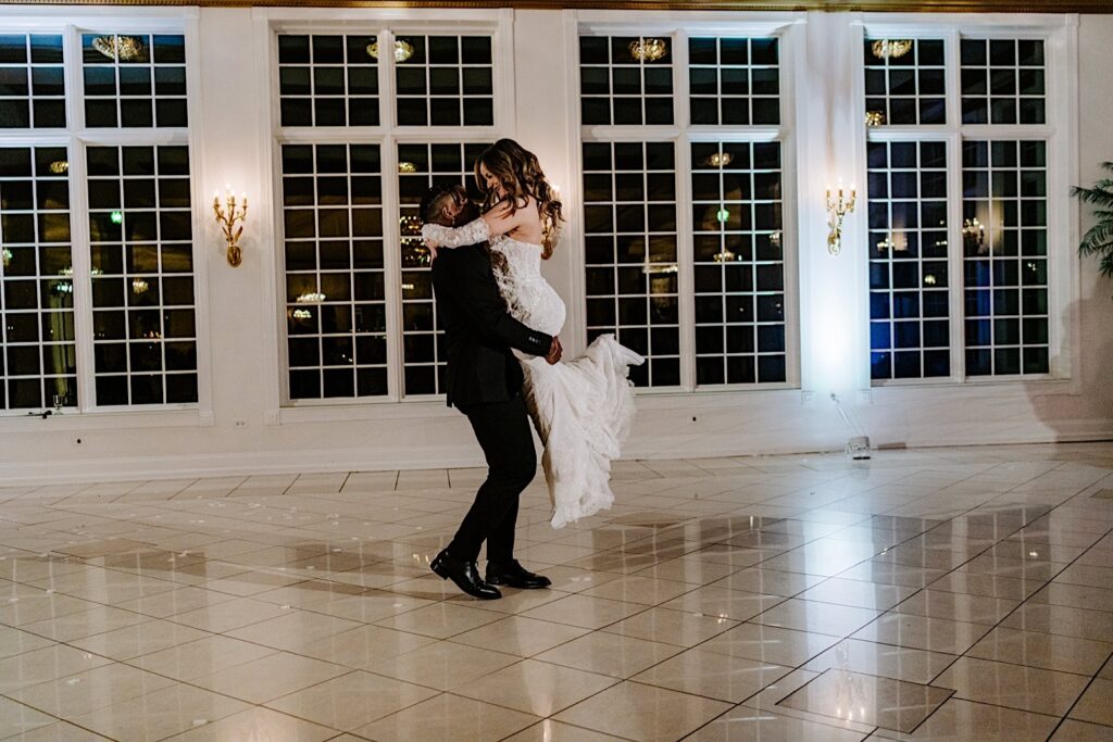 A groom lifts the bride in the air as she smiles at him during their first dance at their indoor wedding reception at The Haley Mansion