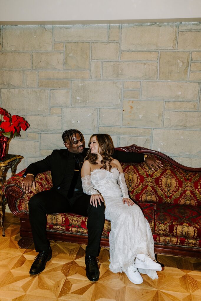 A bride and groom sit together on a couch inside of The Haley Mansion and smile at one another