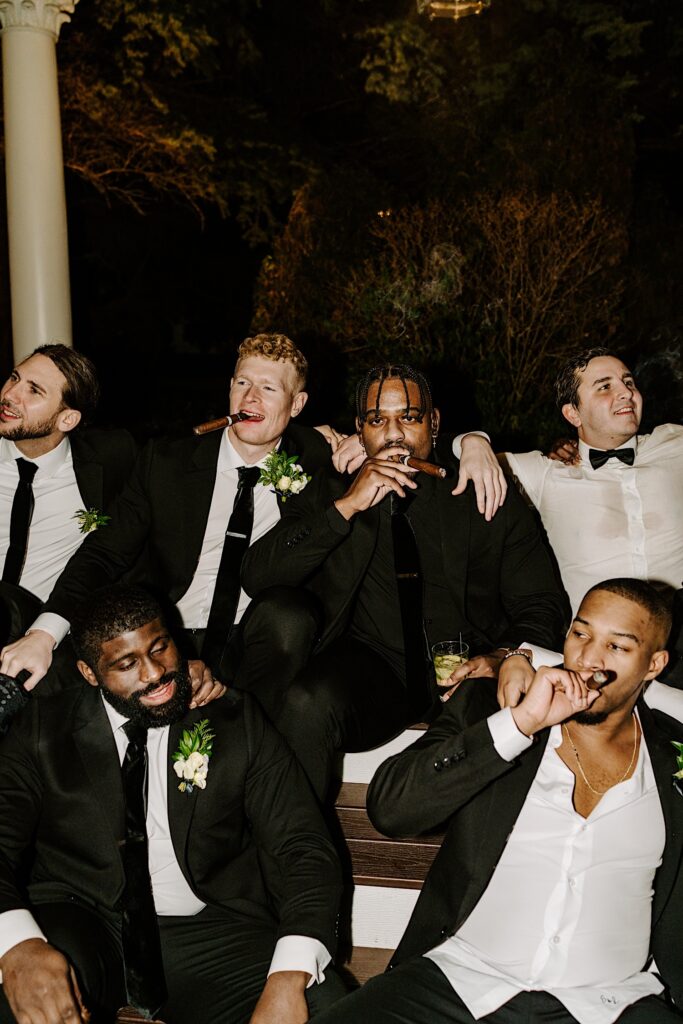 A groom poses with his groomsmen for a photo outside of The Haley Mansion as they all smoke cigars