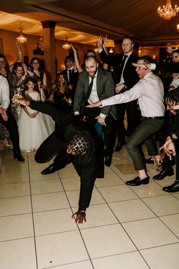 A groom breakdances on the dance floor of his wedding reception as guests around him cheer