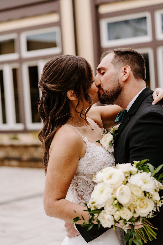 A bride and groom kiss one another while standing outside their wedding venue