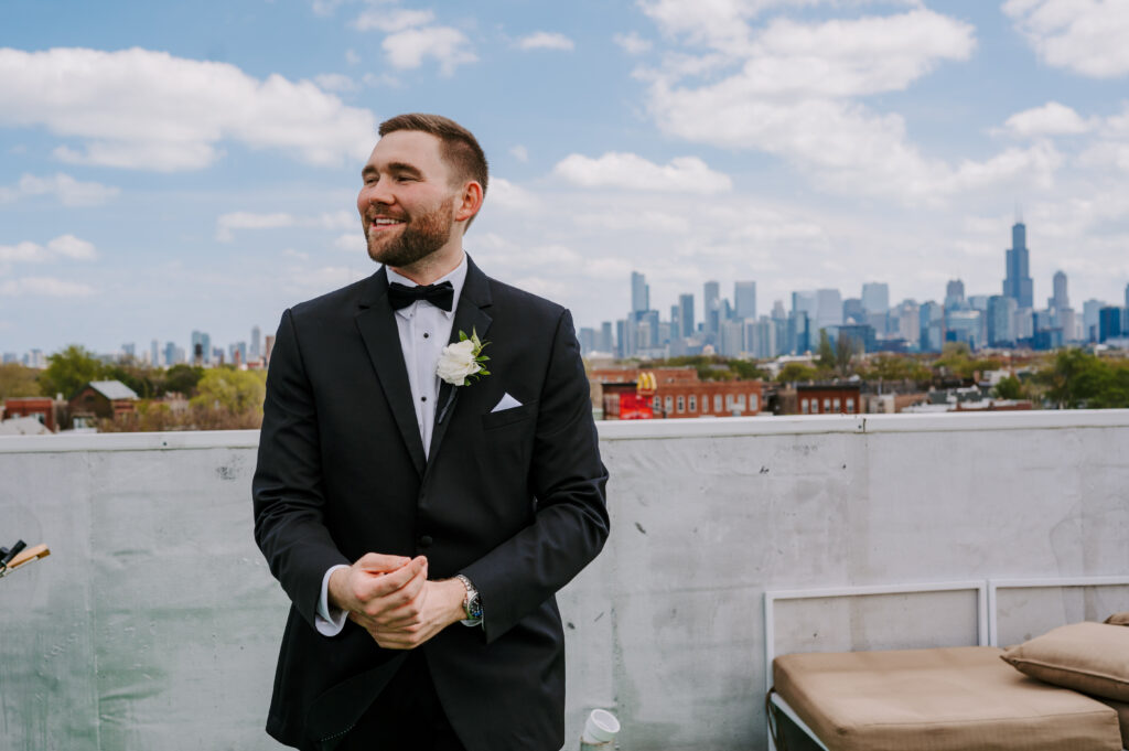 Groom smiles white fixes the cuffs to his black suit on a rooftop that shows the Chicago skyline and a cloudy blue sky behind him. 